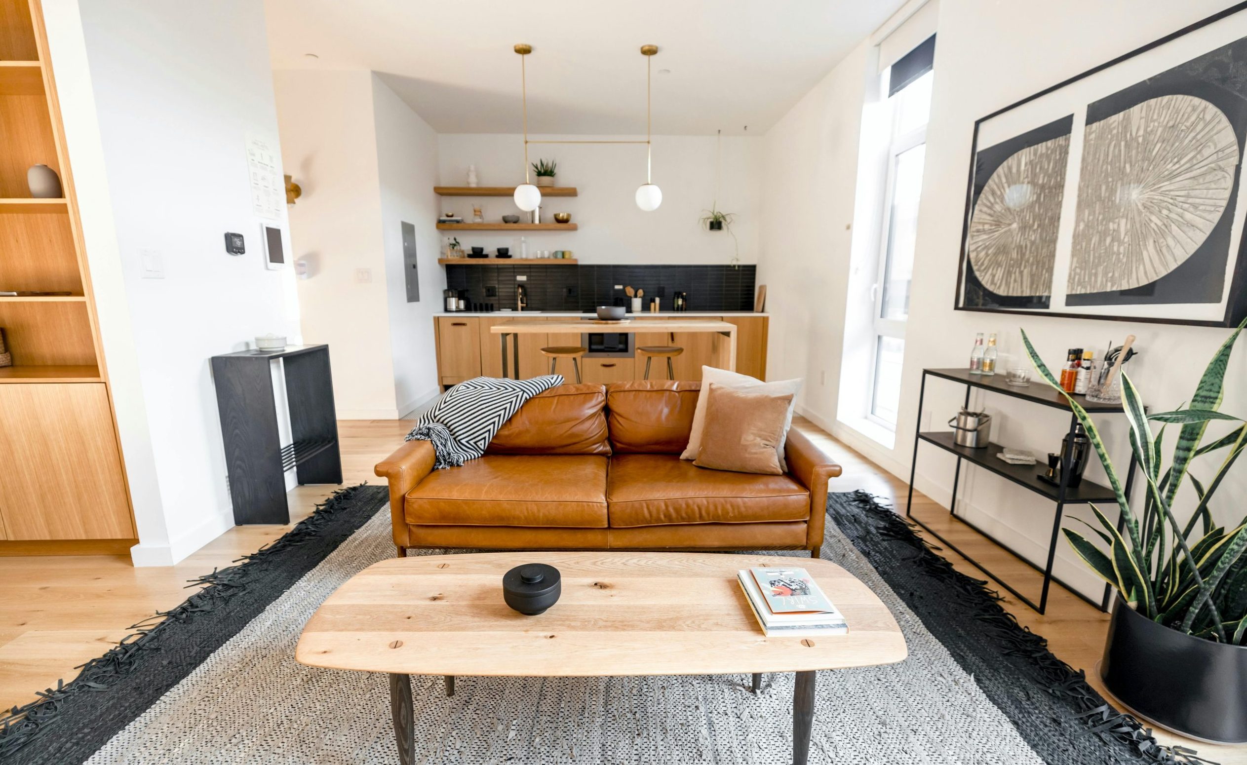 Salon moderne avec canapé en cuir camel, table basse en bois clair, cuisine ouverte en bois avec dosseret noir, grande plante verte et décoration murale minimaliste – intérieur lumineux et accueillant idéal pour un Airbnb.